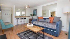 A spacious living room with a blue sofa, a wooden coffee table, and a teal armchair. There is a kitchen area with a bar counter and stools. - Cozy two bedroom, sleeps 6. Wifi, full kitchen, balcony, and pool. (Honolulu)