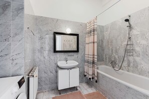 Une salle de bain moderne avec un mur recouvert de carreaux de marbre, un lavabo blanc, un miroir, une douche équipée d’un pommeau de douche à main et un porte-serviettes.