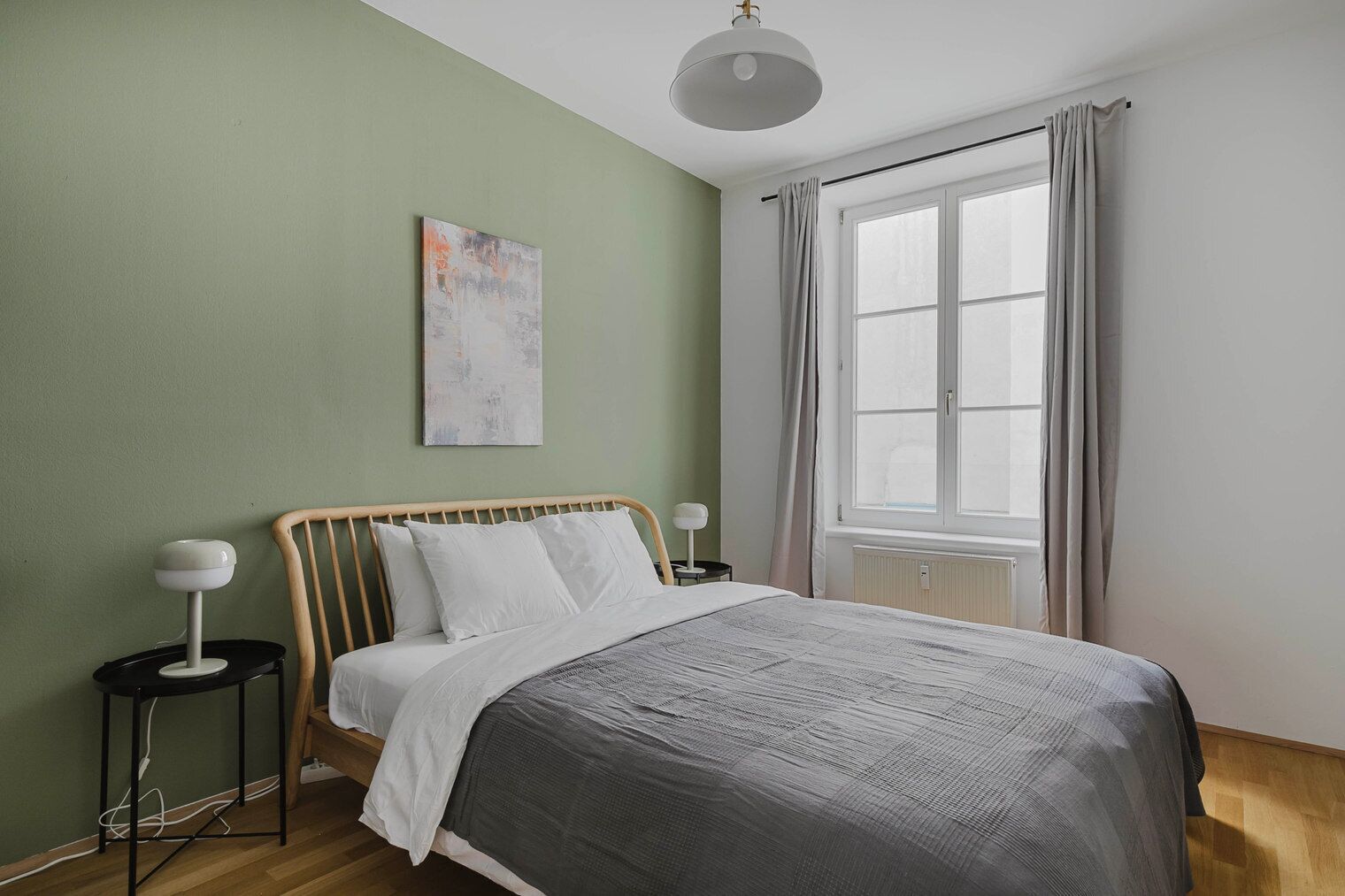 Ein Schlafzimmer mit einem Bett, einem Nachttisch, einer Lampe, einem Fenster mit Vorhängen und einem Bild an der Wand.