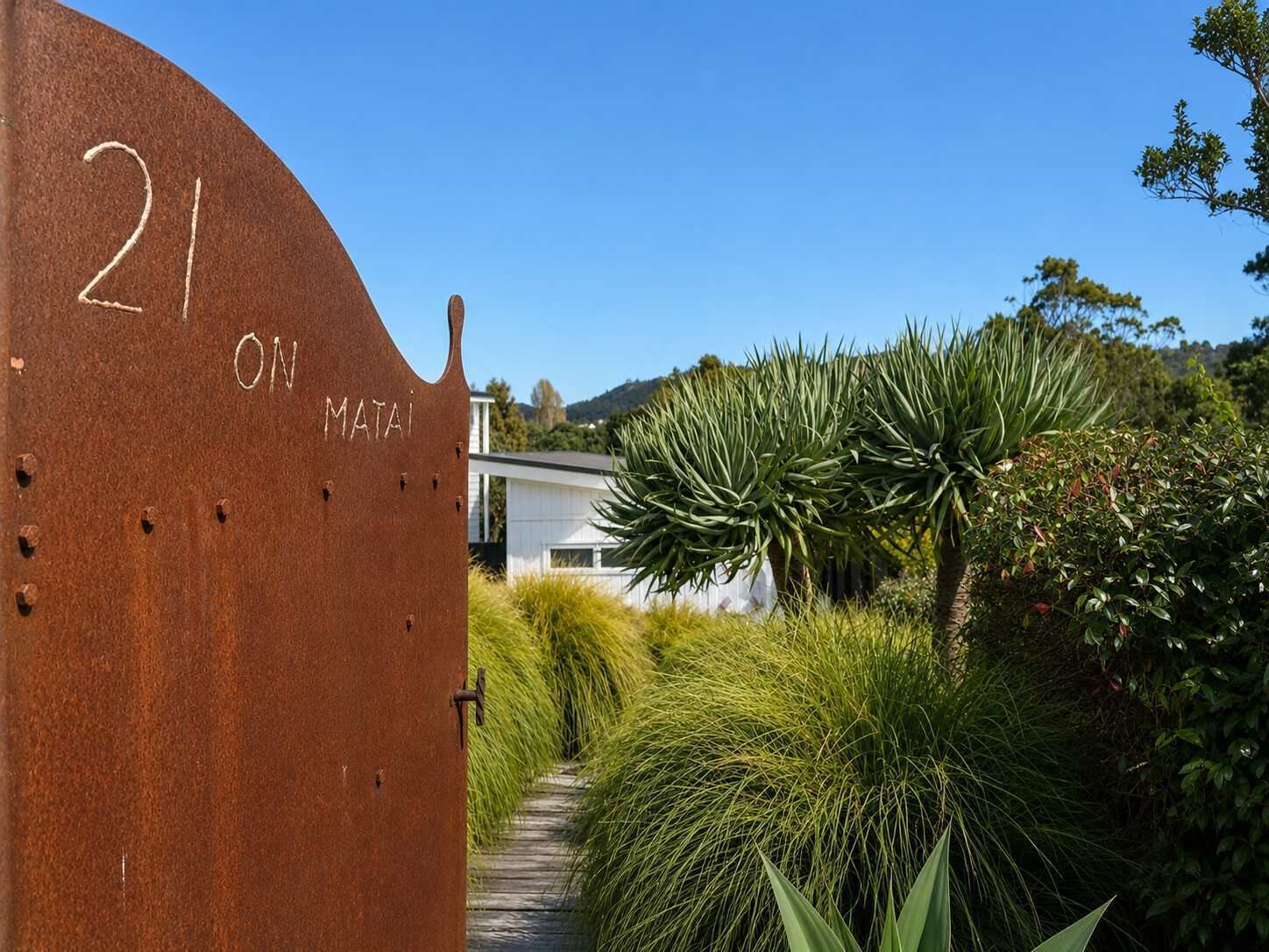 A metal gate with the number 21 and the word 'MATAI' engraved on it, leading to a pathway surrounded by lush greenery and a white building in the background.