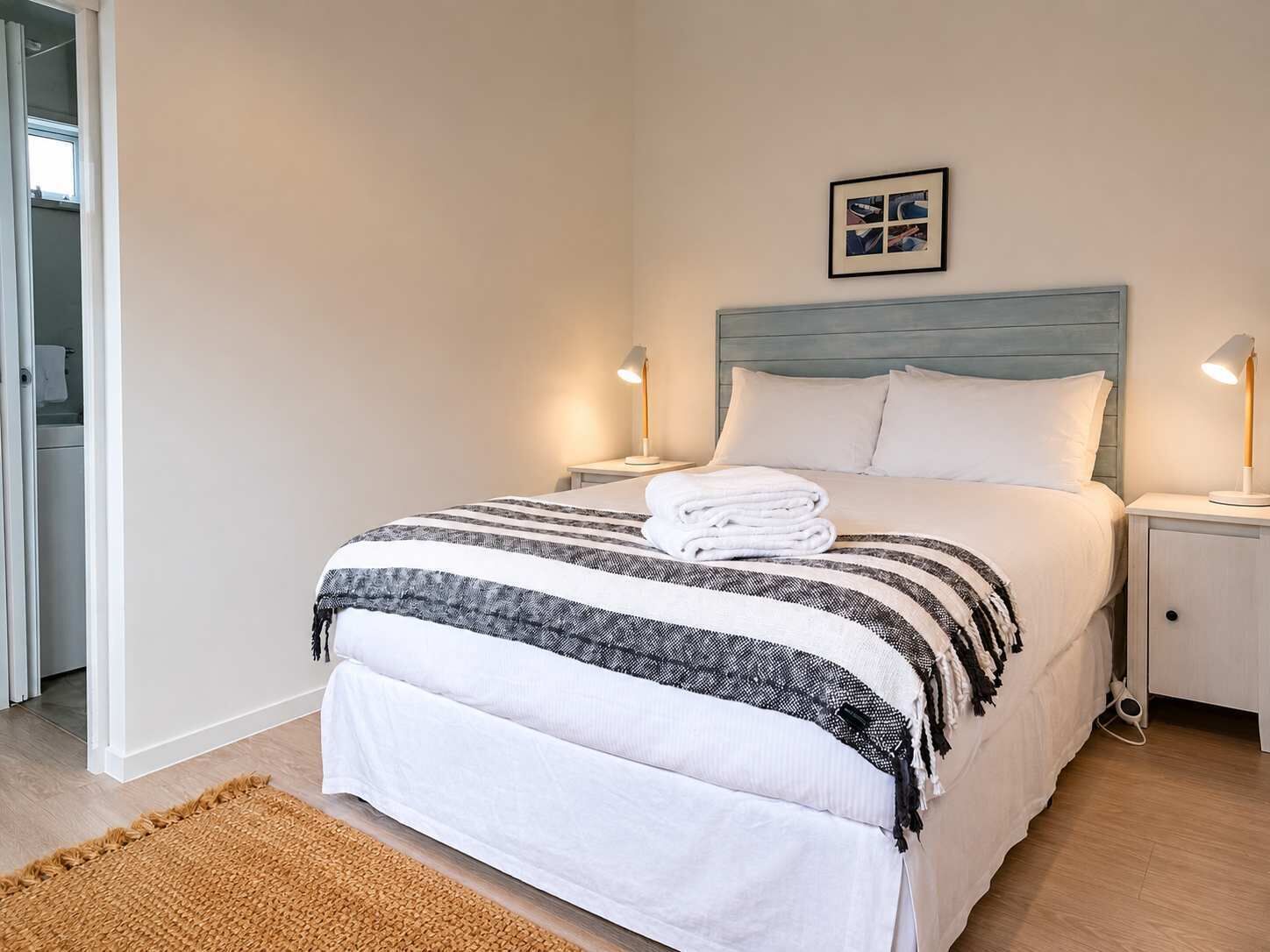 A neatly made bed with a striped blanket and a folded towel, two bedside lamps, a framed picture on the wall, and a woven rug on the floor.