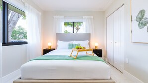 A neatly made bed with white linens, a green throw blanket, and a tray with fruits. There are two bedside tables with lamps, a window with curtains, and a framed plant on the wall. - Welcome to Casa Luna – Your Private Oasis with Heated Pool! (Cutler Bay)
