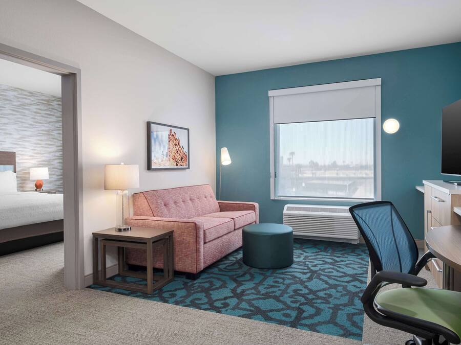 Habitación de hotel con cama, sofá, escritorio, silla, lámpara, un cuadro en la pared y ventana con vistas.