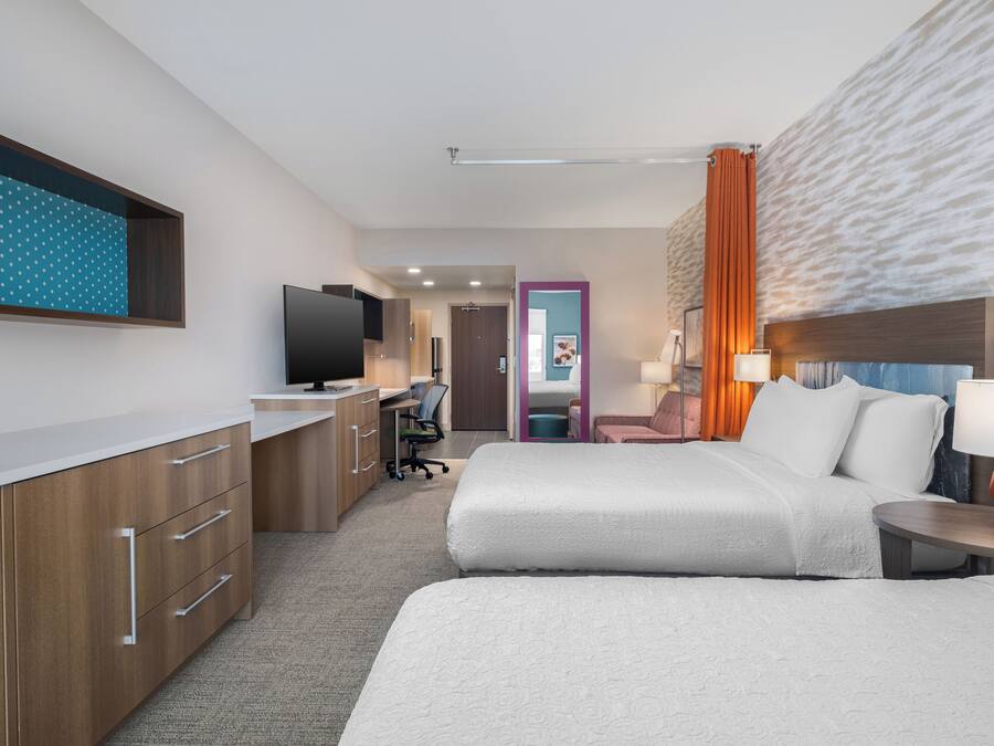 Habitación de hotel con cama, mesita de noche, lámpara, televisor, escritorio, silla y puerta al baño.