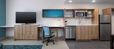 A modern kitchen with a flat-screen TV, wooden cabinets, a microwave, and a stainless steel refrigerator.
