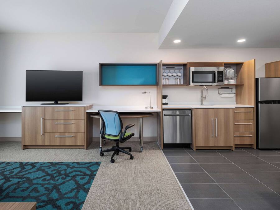 A modern kitchen with a flat-screen TV, wooden cabinets, a microwave, and a stainless steel refrigerator.