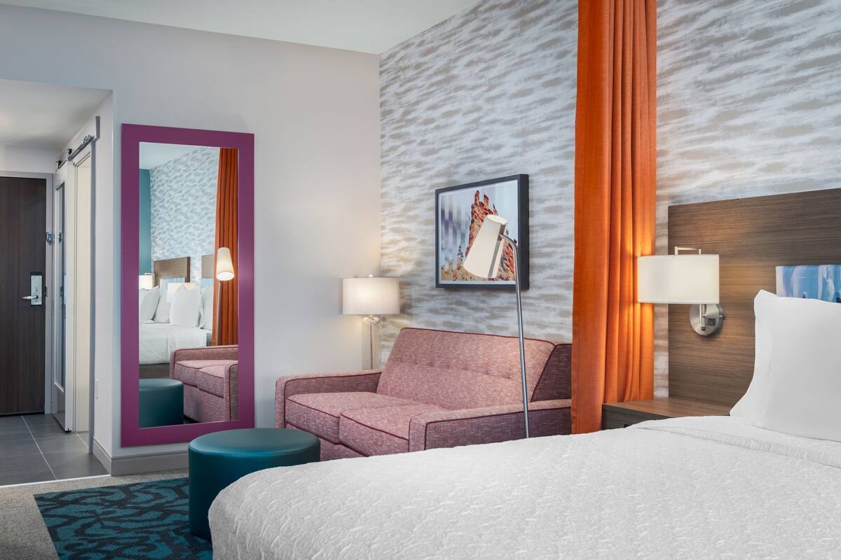 A hotel room with a bed, a pink armchair, a teal ottoman, a large mirror, a wall-mounted lamp, and a framed picture.