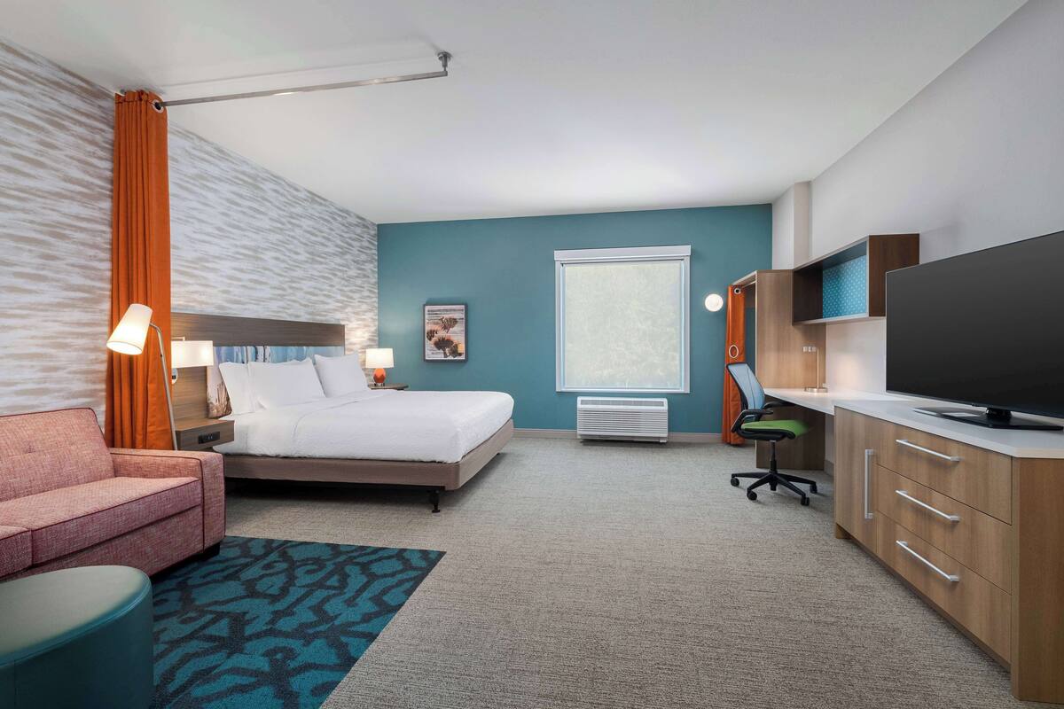 A hotel room with a bed, a desk with a flat-screen TV, a chair, a sofa, and a large window.