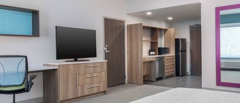 A modern room with a bed, a desk with a computer, a TV, a kitchen area, and a bathroom.