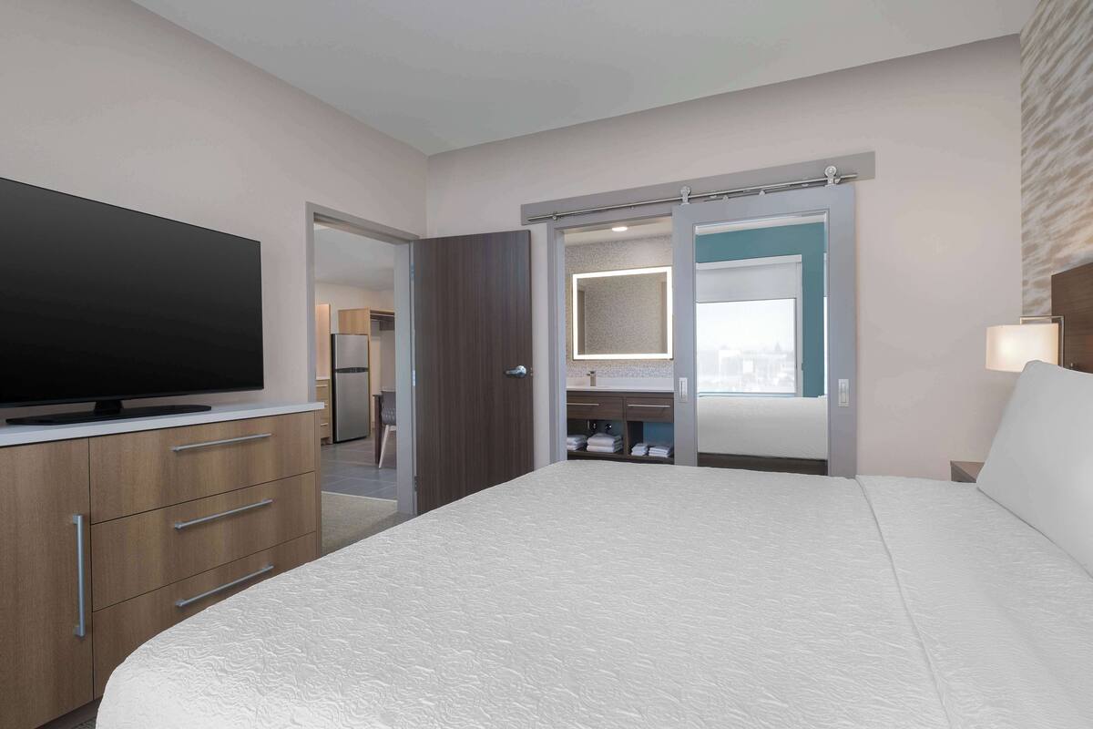 A hotel room with a large bed, a flat-screen TV, wooden cabinets, a bathroom with a mirror, and a sliding door closet.