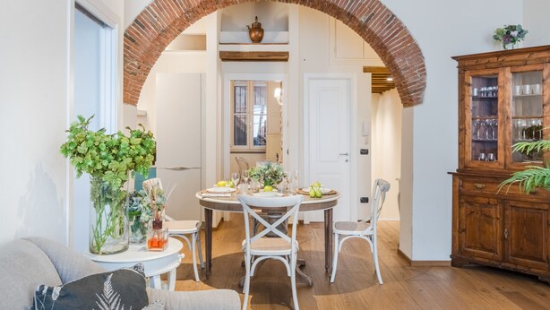 A dining area with a brick archway, white chairs, a round table set with plates, glasses, and a vase of flowers. - Paolino View Apartment (Lucca)