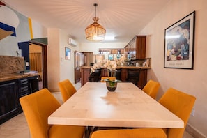 Et spiseområde med et langt bord, orange stole, en hængende lampe og et køkken i baggrunden.
