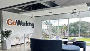 A modern co-working space with a large sign reading "CoWorking", a curved blue sofa, a white table with stools, and a view of a playground through large windows.