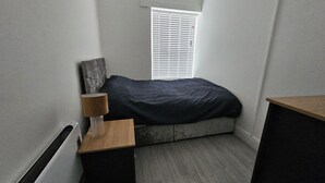 A small, sparsely furnished room with a bed, a nightstand, a lamp, and a window with blinds.