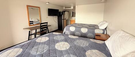 A bedroom with two beds, a desk, a chair, a television, and a kitchenette.
