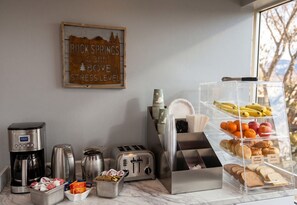 En frukostplats med en kaffebryggare, brödrost och ett bord där frukt och bröd är framdukat.
