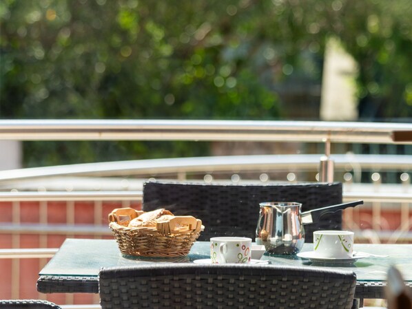 A wicker table with a basket, teapot, and cups, set on a balcony with a railing and greenery in the background.