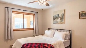 Une chambre à coucher avec un lit, un ventilateur de plafond, une fenêtre avec des rideaux et un tableau au mur.