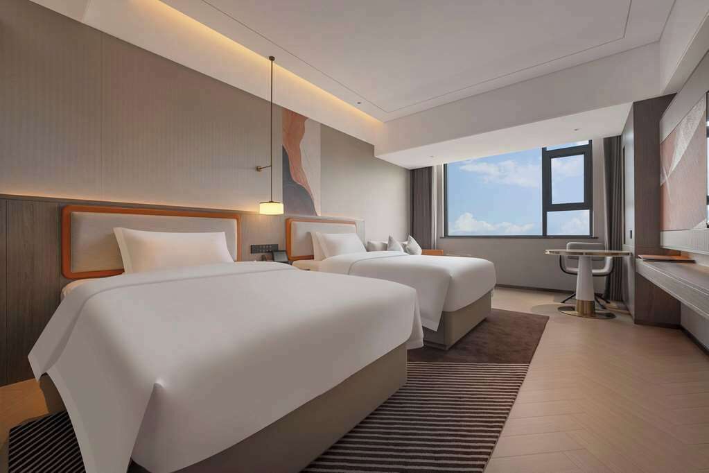 A modern hotel room with two beds, a desk, a chair, and a large window offering a view of the sky.