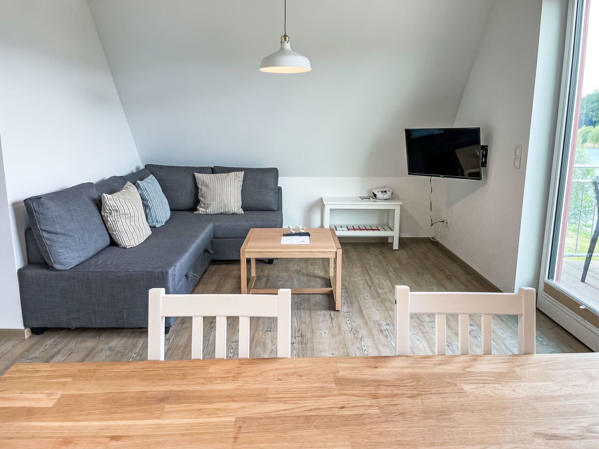 Ein modernes Wohnzimmer mit einem grauen Ecksofa, einem Holztisch, weißen Sesseln, einer weißen TV-Konsole und einem großen Fenster.