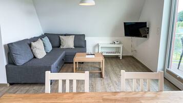 Ein modernes Wohnzimmer mit einem grauen Ecksofa, einem Holztisch, weißen Sesseln, einer weißen TV-Konsole und einem großen Fenster.