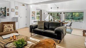 A spacious living room with a black leather sofa, a wooden coffee table, and a dog. There is a dining area with a table and chairs, and a kitchen visible through an open door.
