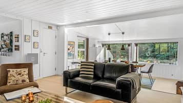 En rummelig stue med en sort lædersofa, et træbord og en hund. Der er et spiseområde med et bord og stole, og et køkken kan ses gennem en åben dør.