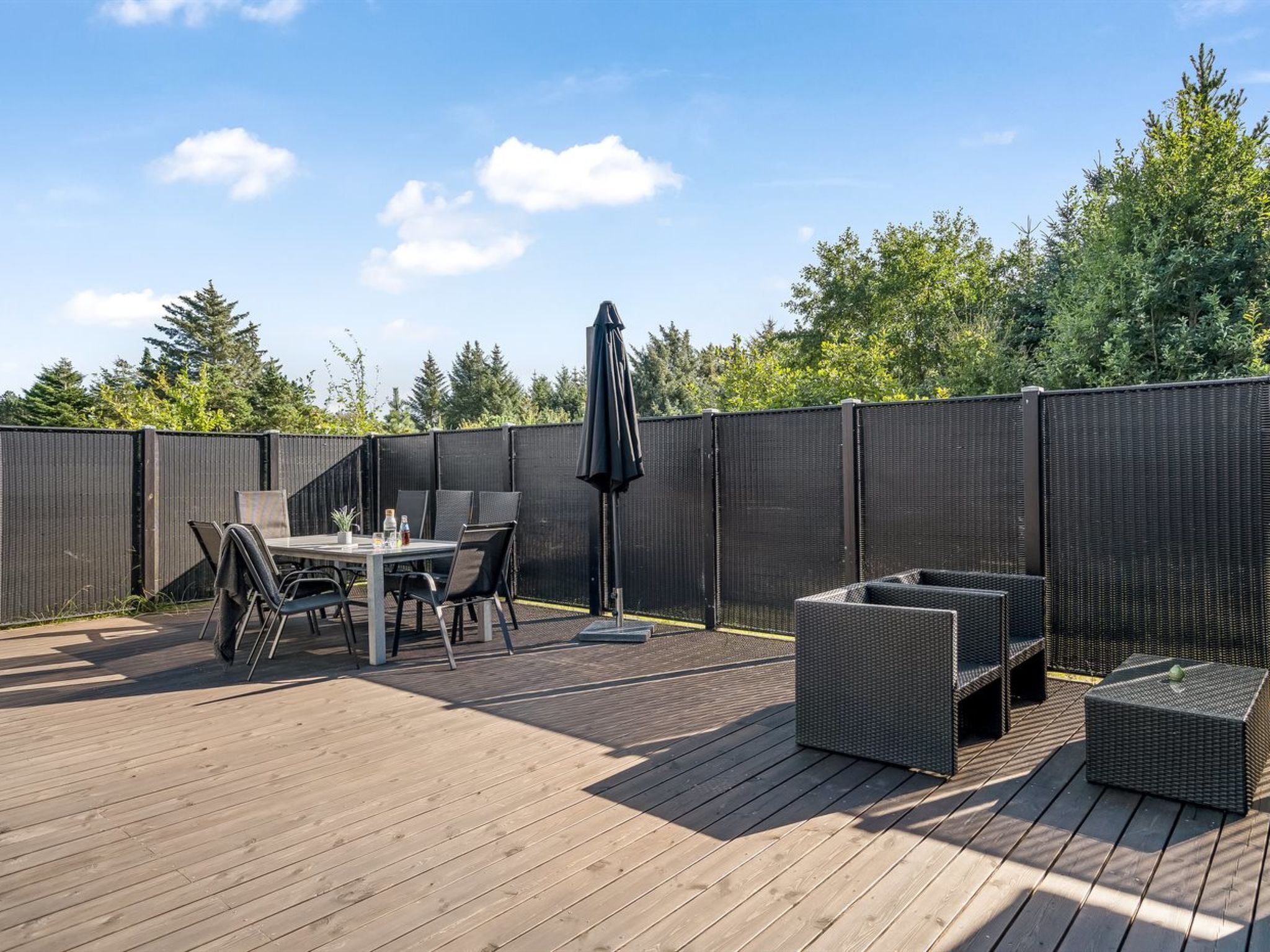 Eine Holzterrasse mit Tisch und Stühlen, einer Korbsitzgruppe und einem großen Sonnenschirm.