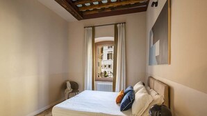 Un dormitorio con una cama, una mesita de noche, una silla y vistas a edificios a través de una ventana.