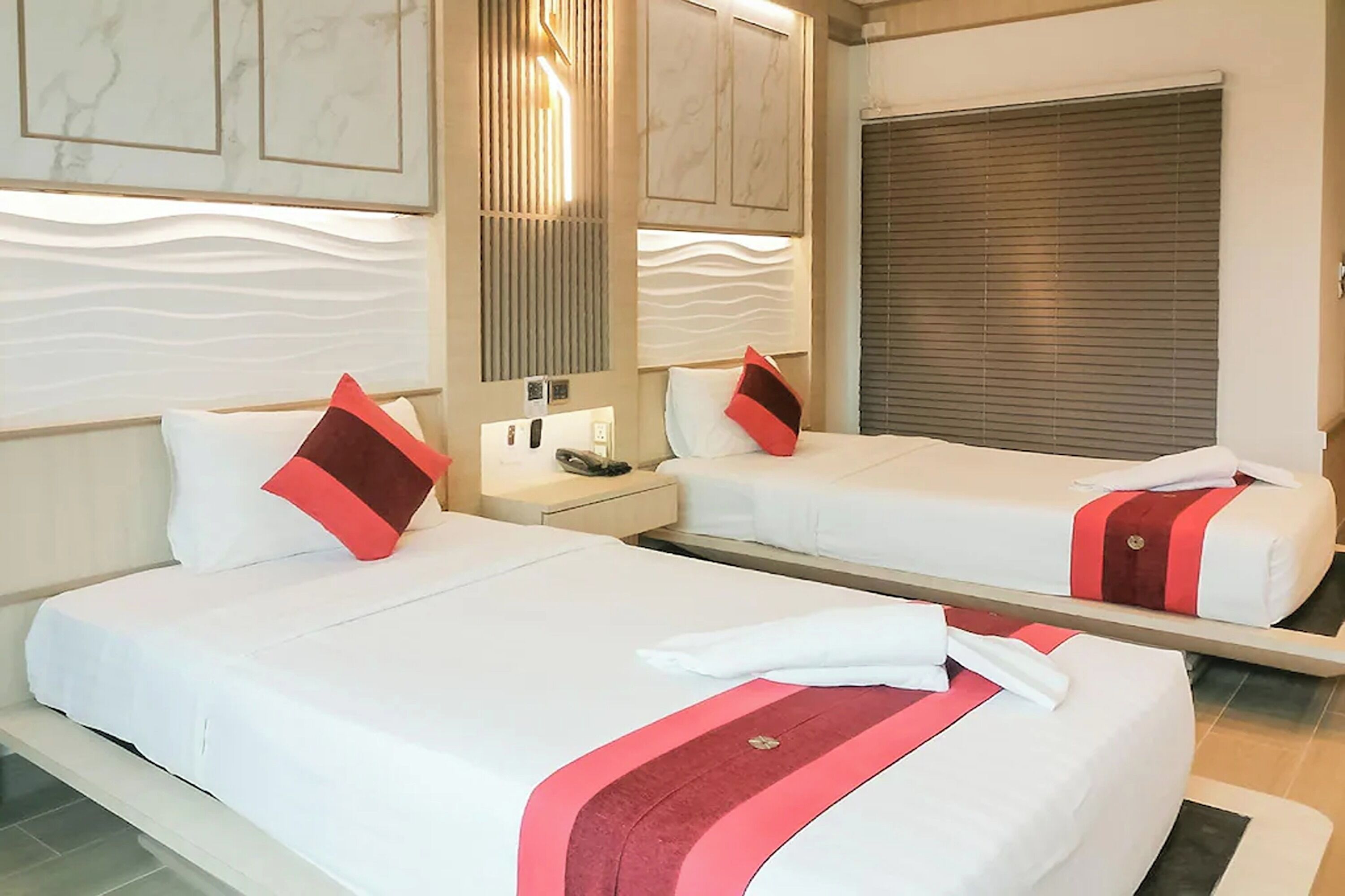 A hotel room with two beds, each with white bedding and red accents, a nightstand with a phone, and a large window with blinds.