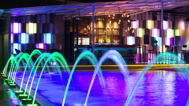 A pool with illuminated water jets and a bar area with colorful lighting in the background. - Ruean Phae Royal Park Hotel (Phitsanulok)