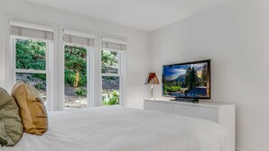 A bedroom with a large bed, a television on a white console, and a window with a view of greenery. - Palisades Condo – Walk to Ski Lifts, Mtn Views (Tahoe City)