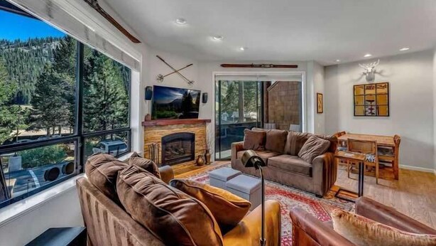 A modern living room with a fireplace, large windows, and a view of a forest. - Palisades Condo – Walk to Ski Lifts, Mtn Views (Tahoe City)