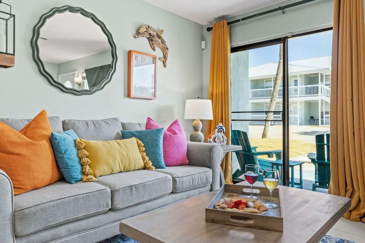 A living room with a grey sofa, colorful cushions, a wooden coffee table with snacks and drinks, a large mirror, a lamp, and a view of a pool and building through large windows.