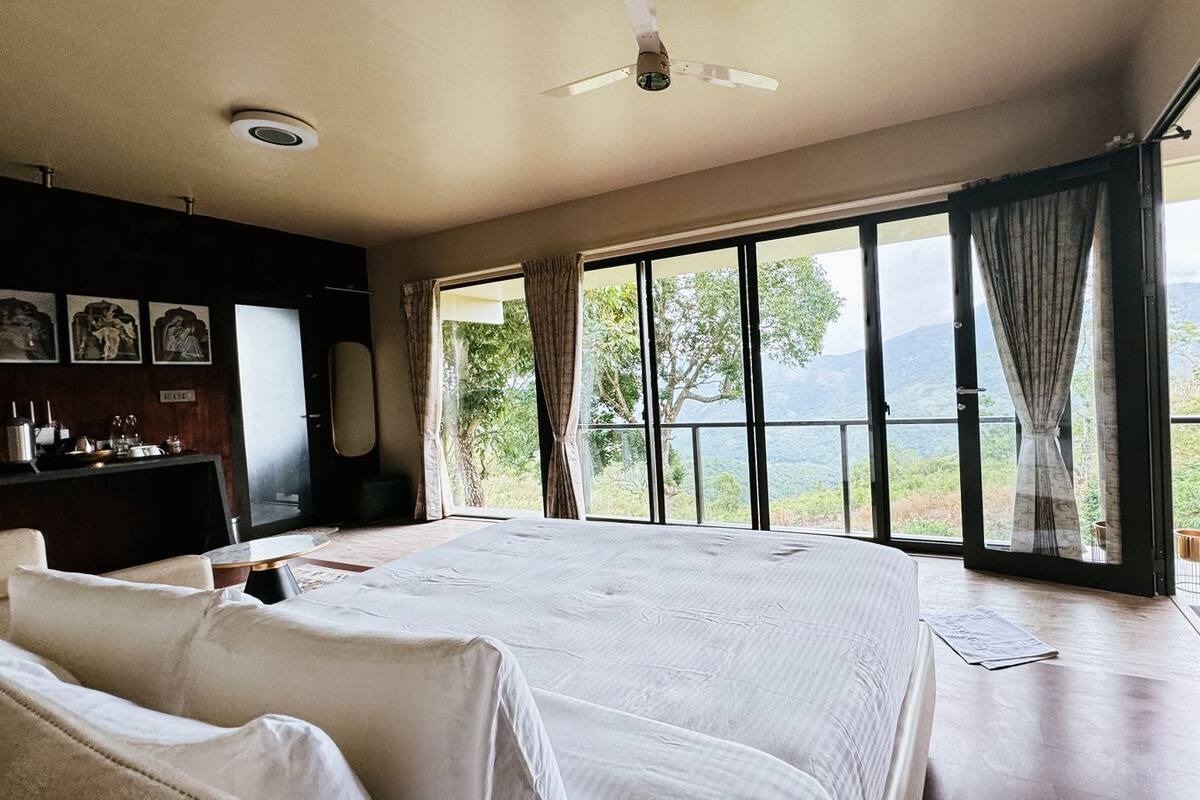 Quarto com uma cama grande, uma escrivaninha com cadeira e vista para montanhas por grandes janelas.