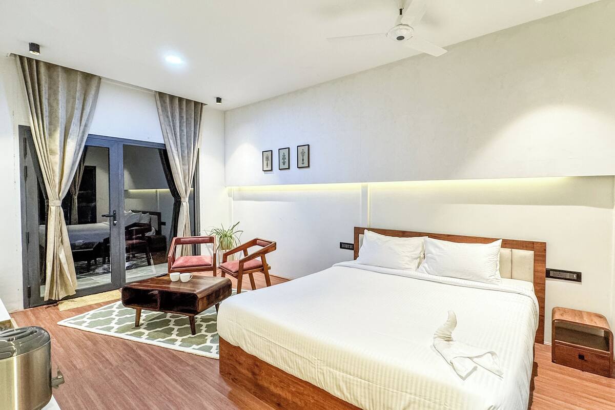 Quarto de hotel com uma cama grande, uma escrivaninha com cadeira, uma pequena mesa com duas chávenas, uma planta em vaso e vista para o exterior através de uma porta de vidro.