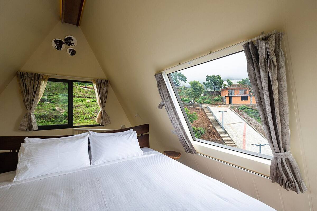 Quarto com cama, duas janelas e vista para um jardim e edifícios lá fora.