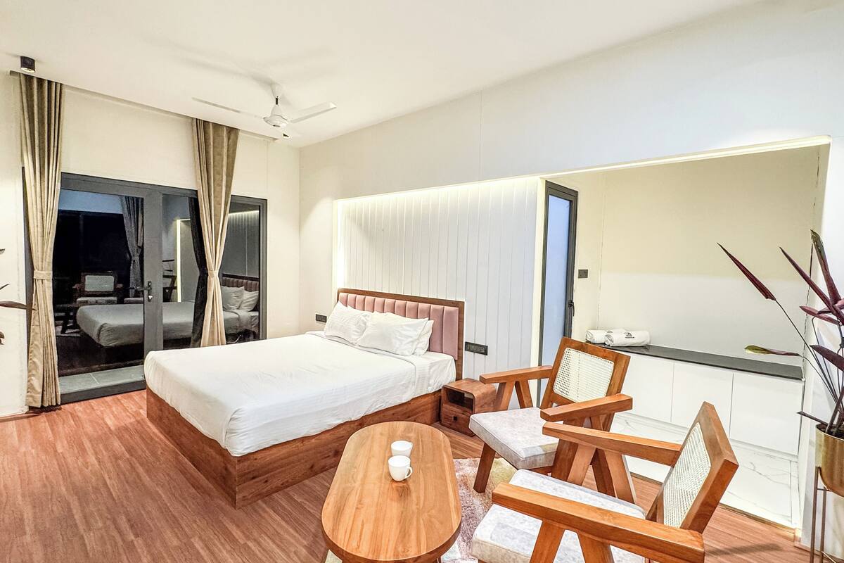 Quarto de hotel com cama, mesa de madeira, duas cadeiras, ventoinha no teto e uma janela grande.