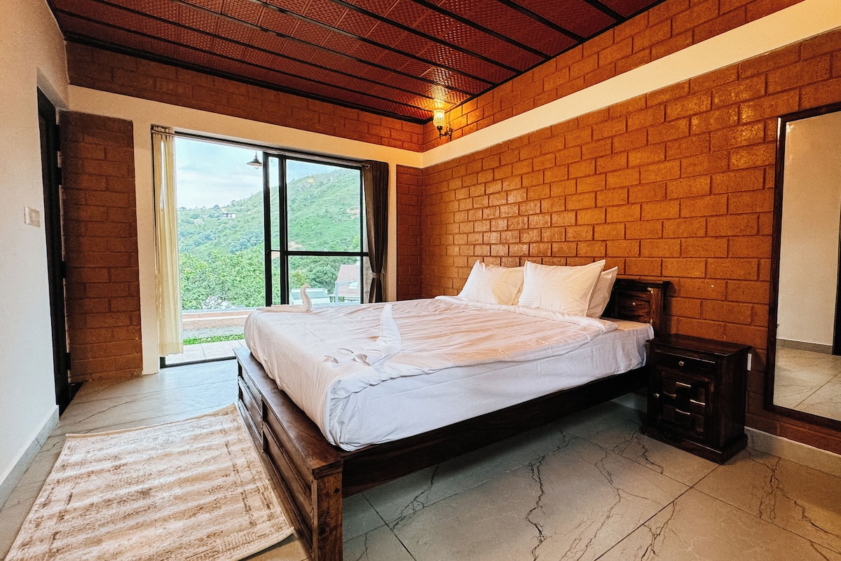 Quarto com cama grande, mesa de cabeceira, espelho e vista para o verde pela janela.