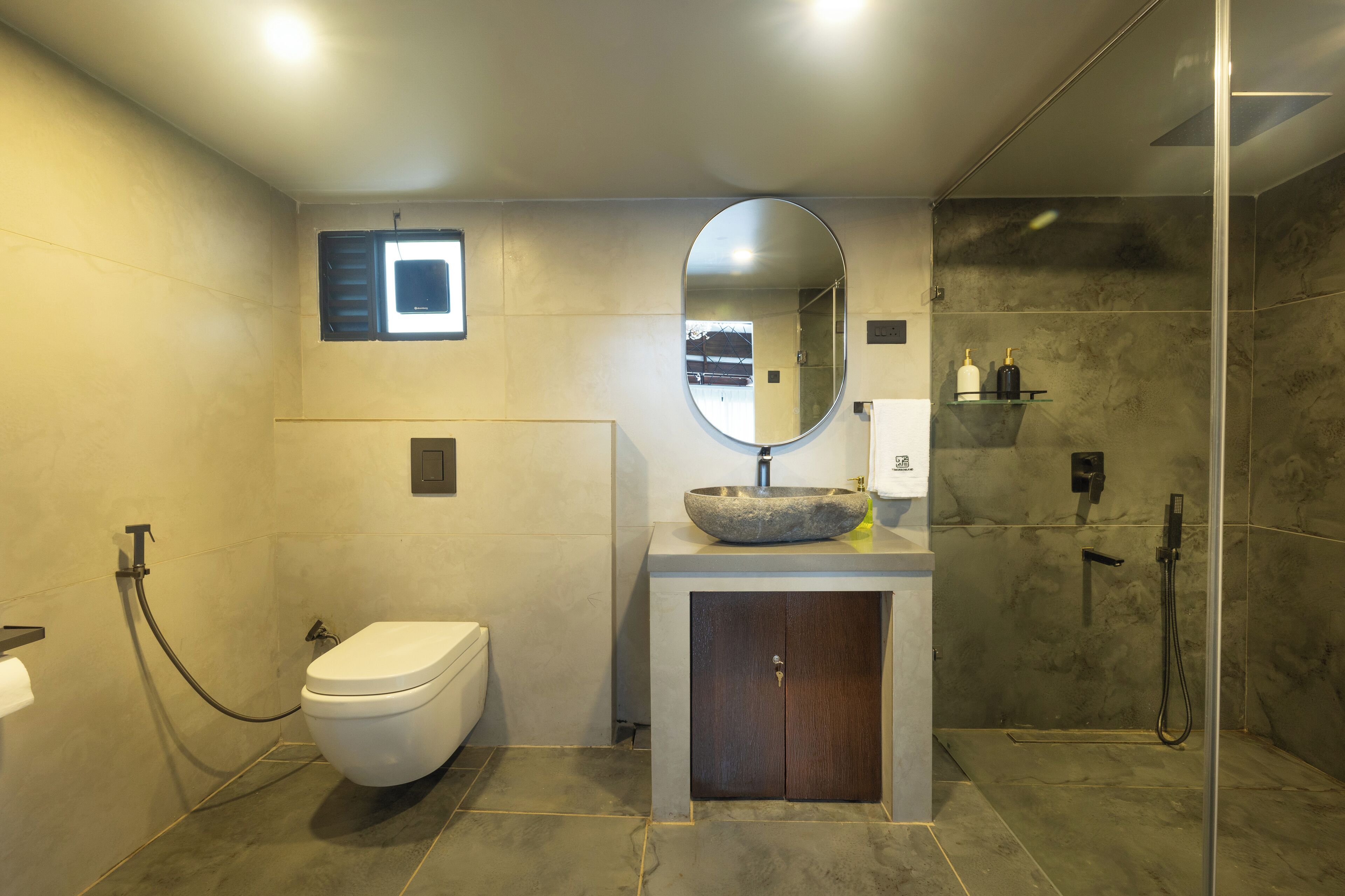 A modern bathroom with a toilet, a large oval mirror, a stone sink, and a glass-enclosed shower area.