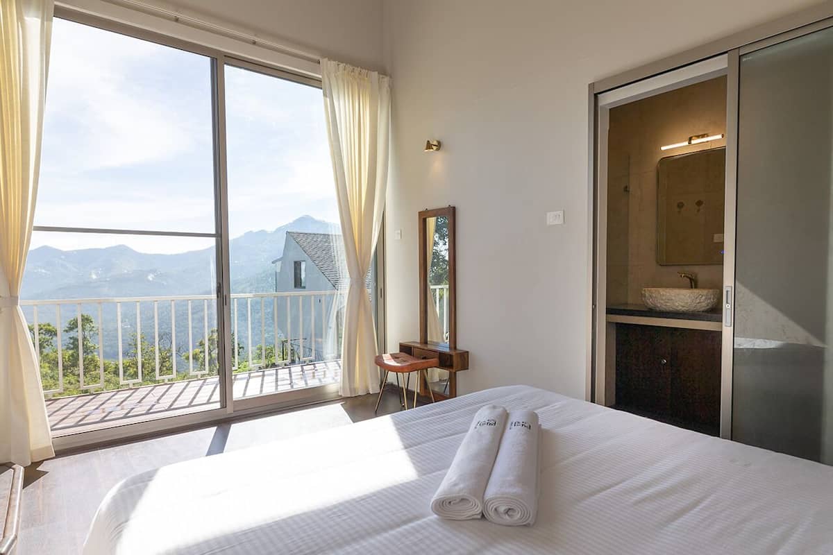 Quarto de hotel com uma cama grande, banheiro com espelho e pia, e varanda com vista para as montanhas.