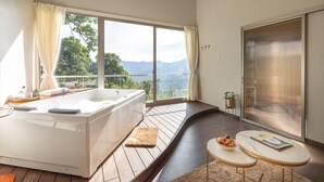 A modern bedroom with a large window, a bathtub, a small table with a bowl of fruit, and a view of mountains. - TOMORROWLAND  FARMS AND SUITES (Kodaikanal)