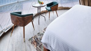 A room with a bed, two chairs, a small table with fruit, and a view of mountains through large windows. - TOMORROWLAND  FARMS AND SUITES (Kodaikanal)