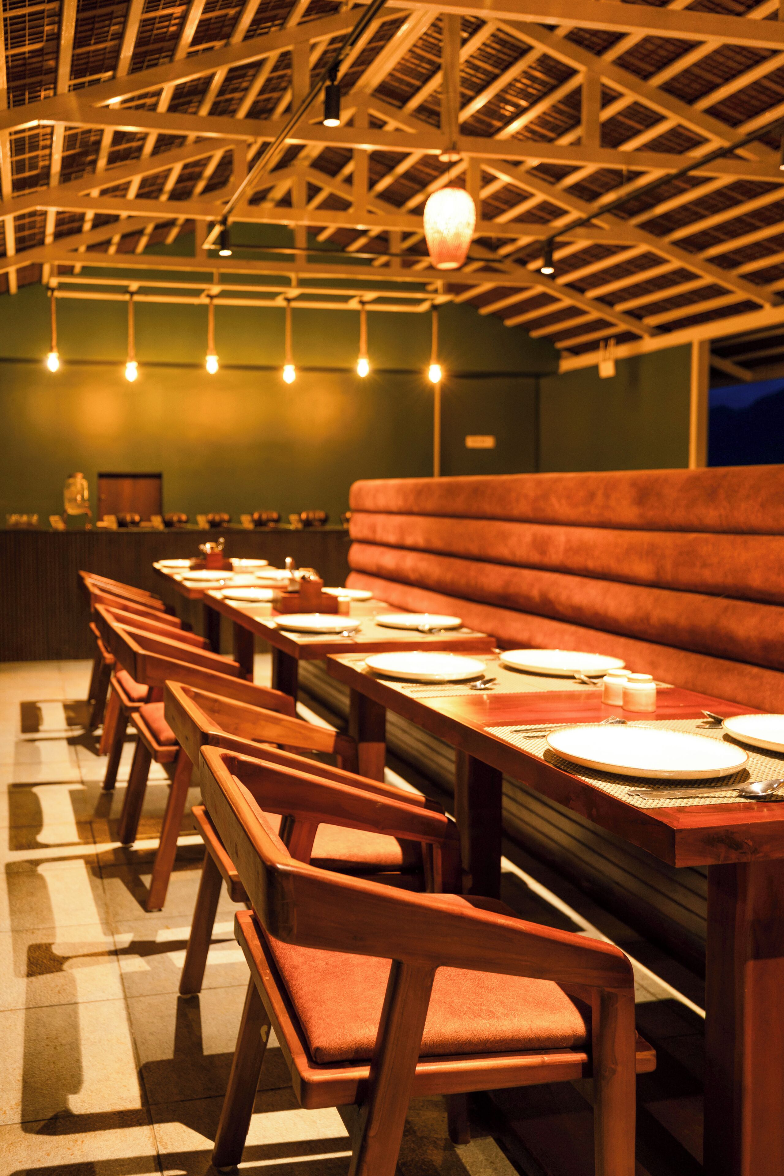 Un restaurante con mesas y sillas de madera, un techo alto con vigas a la vista y lámparas colgantes.