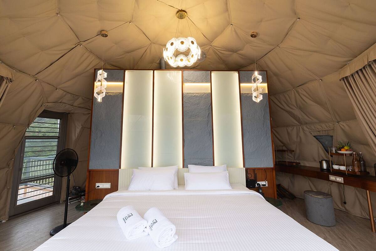 Quarto tipo tenda com cama, mesas de cabeceira, ventilador e luminária no teto.
