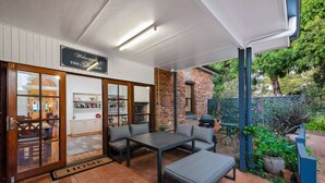 Un patio techado con mesa y asientos, un muro de ladrillo y un área de jardín con plantas y una parrilla.