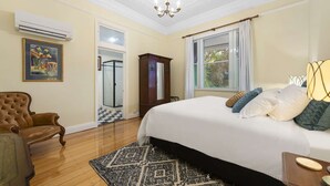 Un dormitorio con una cama grande, una silla de madera, un escritorio, una araña y una ventana con cortinas.