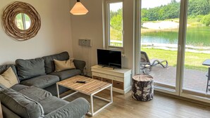 Een moderne woonkamer met een grijze bank, een houten salontafel, een televisie op een witte standaard en een groot raam met uitzicht op een meer en begroening.