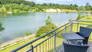 A deck with a table and chairs overlooking a lake, surrounded by greenery and a forested hillside.