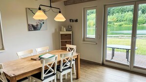 A dining area with a wooden table and white chairs, a pendant light, a painting, and a view of a green landscape through a glass door.
